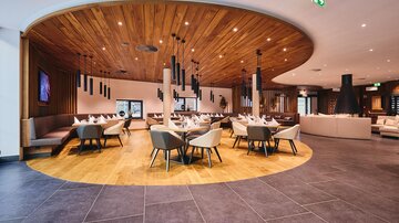A modern restaurant with a wood-paneled ceiling and stylish seating areas. The decor is bright and inviting, with round tables and elegant chairs.