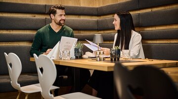 A cozy café with a wooden-paneled atmosphere. Two people are sitting at a table and conversing while looking at the menus. | © Silvrettaseilbahn AG