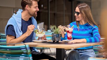 A couple sits at an outdoor table enjoying desserts and cocktails. The scene is bright and inviting, with cheerful weather.