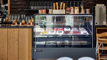 An ice cream counter in a modern café with various ice cream flavors. In the background, glasses and waffles are elegantly arranged on a shelf.