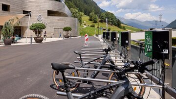 A modern bike station with e-bikes is in front of an elegant building. In the background, there are green hills and a blue sky.