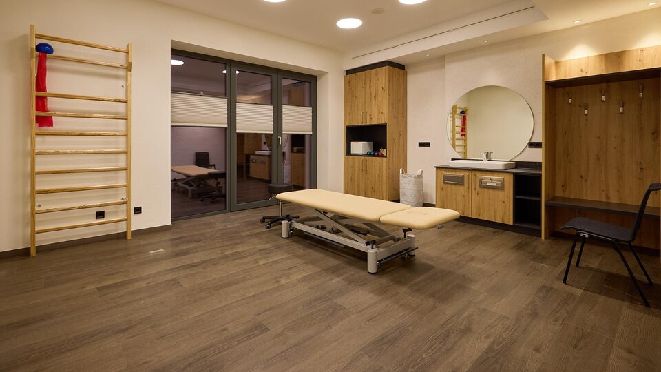 A bright, modern therapy room with a treatment table and a wall with various training equipment. There is a mirror and wooden shelves that create an inviting atmosphere.