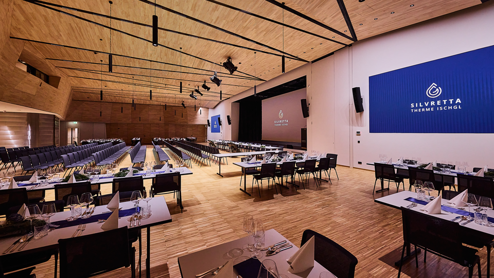 A modern event room with several tables elegantly set. The room has a large screen and wooden paneling on the walls.