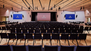 A large, modern event space with many chairs arranged in rows. In the background, there are stage walls with digital displays.
