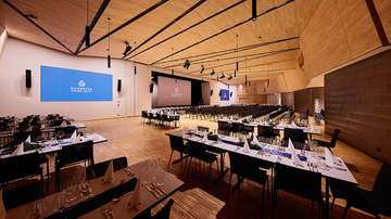 A modernly furnished event space with tables and chairs for a dinner event. Large screens for presentations are visible on the walls.