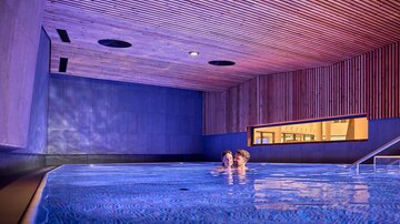 Ein modernes Schwimmbad mit sanftem Licht und Holzverkleidung. Zwei Personen genießen die entspannende Atmosphäre im Wasser.
