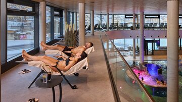 Ein entspannender Wellnessbereich mit zwei Personen auf Liegen. Im Hintergrund ist ein Pool mit farbigem Licht und eine moderne Architektur zu sehen.