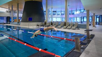 Ein moderner Innenpool mit einem Schwimmer, der ins Wasser springt. Die Umgebung ist hell und einladend, mit Liegen und großen Fenstern.