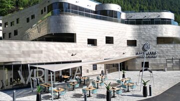 Ein modernes Gebäude mit einer einladenden Terrasse und Tischen. Im Hintergrund sind grüne Berge sichtbar.