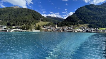 Eine malerische Landschaft mit einem klaren blauen Wasserbecken und Bergen im Hintergrund. Der Himmel ist blau mit einigen weißen Wolken.
