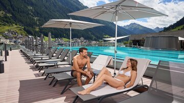 Ein entspannter Tag am Pool mit Liegen und Sonnenschirmen. Ein Paar genießt die Sonne und liest ein Buch.