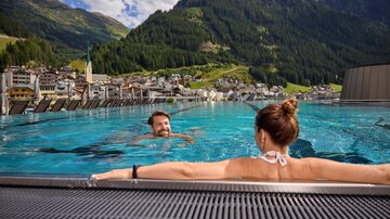 Ein schöner Infinity-Pool mit Blick auf die Berge und ein Dorf im Tal. Zwei Menschen genießen das Wasser und die atemberaubende Landschaft.