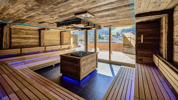 Ein moderner Wellnessbereich mit Holzverkleidung und gemütlichen Sitzgelegenheiten. Große Fenster bieten einen Blick ins Freie und sorgen für viel Tageslicht.