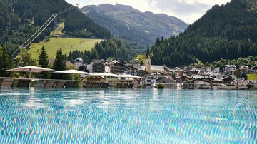 Ein wunderschöner Ausblick auf ein Bergdorf mit grünen Hügeln im Hintergrund. Im Vordergrund ist ein glitzerndes, blaues Infinity-Poolwasser zu sehen.