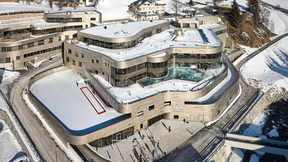 Ein modernes Gebäude in einer schneebedeckten Landschaft mit einem ganzjährig zugänglichen Eisfeld. Die Architektur ist elegant, umgeben von malerischen Bergen und Ferienhäusern.