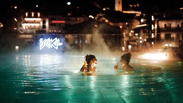 Zwei Frauen genießen drinks im beleuchteten Schwimmbecken bei Nacht. Die Atmosphäre ist entspannt und die Stadtlichter glänzen im Hintergrund.