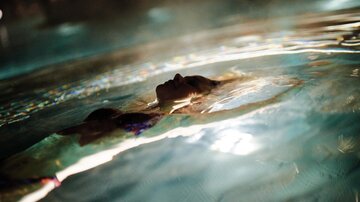 Eine Person schwimmt entspannt im Wasser und blickt nach oben. Die sanften Wellen reflektieren das Licht.