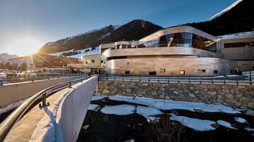 Ein modernes Gebäude in einer alpinen Umgebung. Die Sonne scheint über den Bergen und reflektiert auf der Fassade.