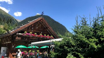 Ein charmantes Holzhaus mit bunten Blumenkästen. Im Vordergrund sitzen Menschen unter Sonnenschirmen in einer malerischen Berglandschaft.
