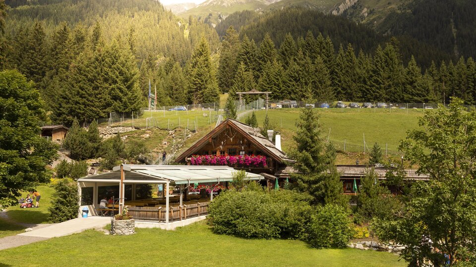 Ein gemütliches Bergrestaurant umgeben von grünen Wiesen und hohen Bäumen. Im Hintergrund sind majestätische Berge und klare Himmel zu sehen.