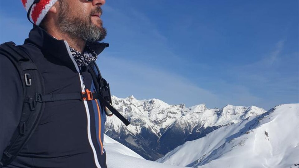 A man with sunglasses and a knit hat stands on a snow-covered mountain peak. In the background, majestic, snow-covered mountains can be seen under a clear blue sky.