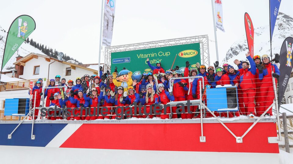 A large group of people in red and blue ski suits is standing on a stage. They are celebrating the Vitamin Cup in a wintry mountain landscape.