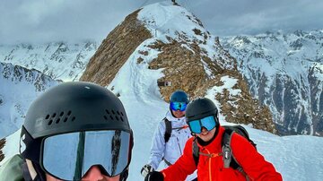 A group of skiers stands on a snow-covered peak. In the background, mountains and a cross are visible.