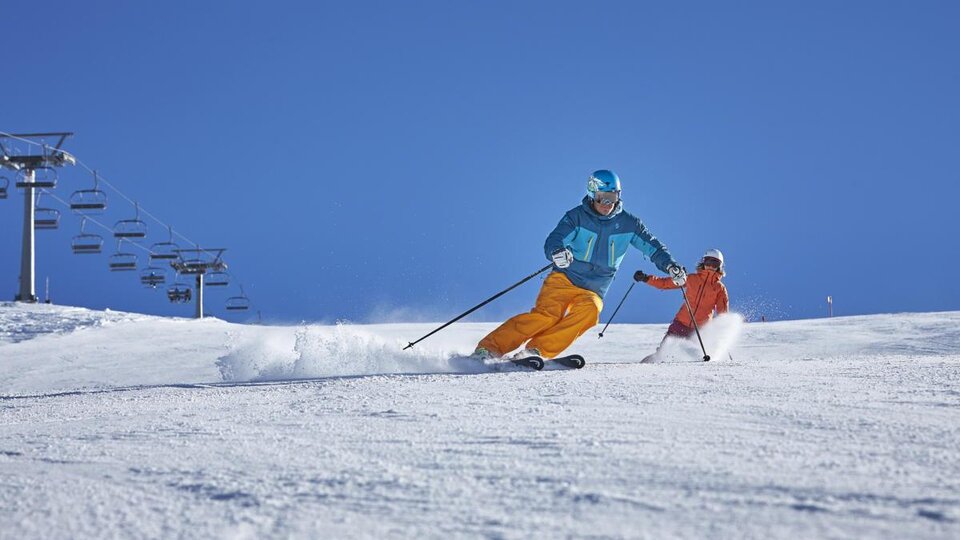 Ein Skifahrer fährt dynamisch den Hang hinunter, während ein Kind in orangefarbener Skibekleidung ihm folgt. Der Himmel ist strahlend blau und die Piste ist mit frischem Schnee bedeckt.