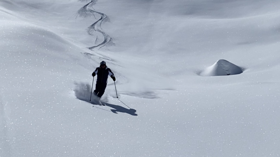 A skier walks through deep, sparkling snow under a clear blue sky. In the background, gentle, snow-covered hills are visible.
