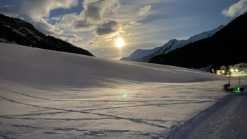 A snowy landscape at night with a bright moonlight. The mountains and the sky are softly illuminated, while the tracks in the snow are visible.