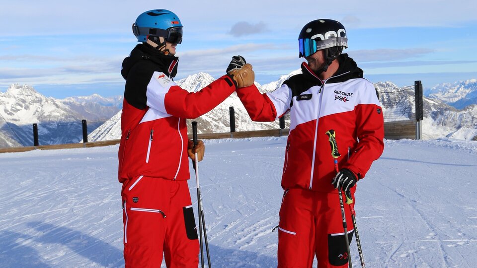 Zwei Skifahrer in roten Skianzügen stehen auf einem schneebedeckten Gipfel. Im Hintergrund sind majestätische Berge zu sehen.