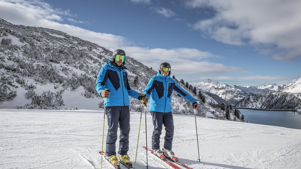 Two skiers are standing on a snow-covered slope. In the background, mountains and a clear sky are visible.
