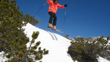 A skier joyfully jumps over a snowy slope. In the background, snow-covered mountains and blue sky can be seen.