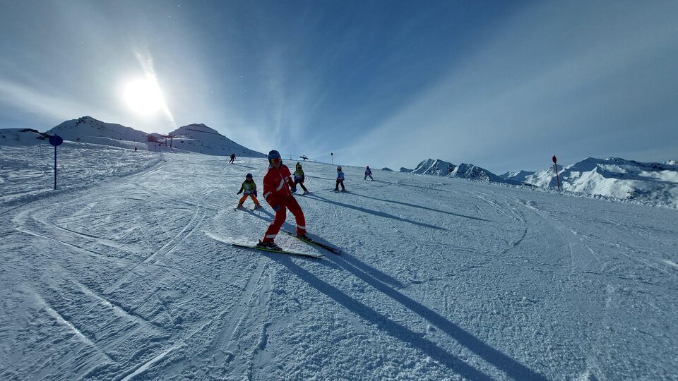 A group of skiing individuals on a sunny slope. Snow-covered mountains can be seen in the background.