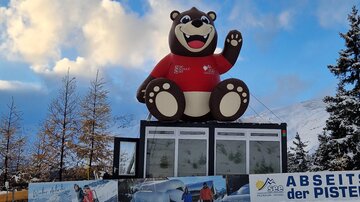 A large, cheerful bear made of plastic sits on a pedestal in the snow. In the background, snow-covered trees and dark clouds can be seen.