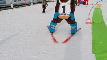 A ski mascot stands on skis in the snow. In the background, children and ski instructors can be seen.