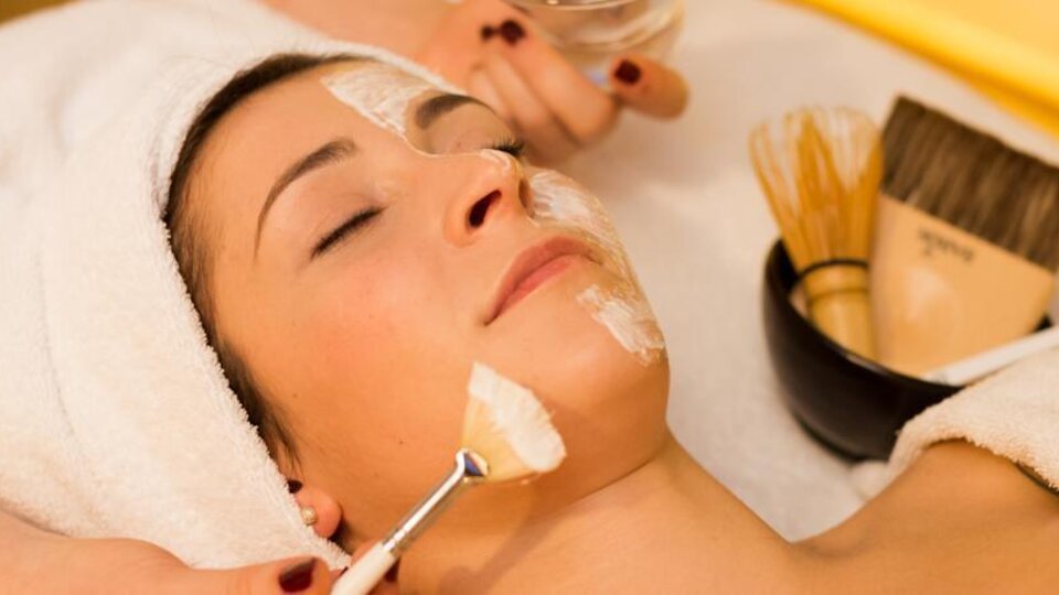 A woman is receiving a facial treatment with a mask on her face. Various beauty tools and a bowl are visible around her.