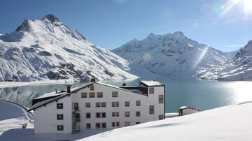 A snow-covered mountain panorama with a tranquil lake in the foreground. In the foreground stands a modern building with large windows.