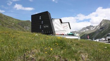 A modern building in the mountains, surrounded by green meadows and blooming flowers. In the background, majestic mountains and a blue sky can be seen.