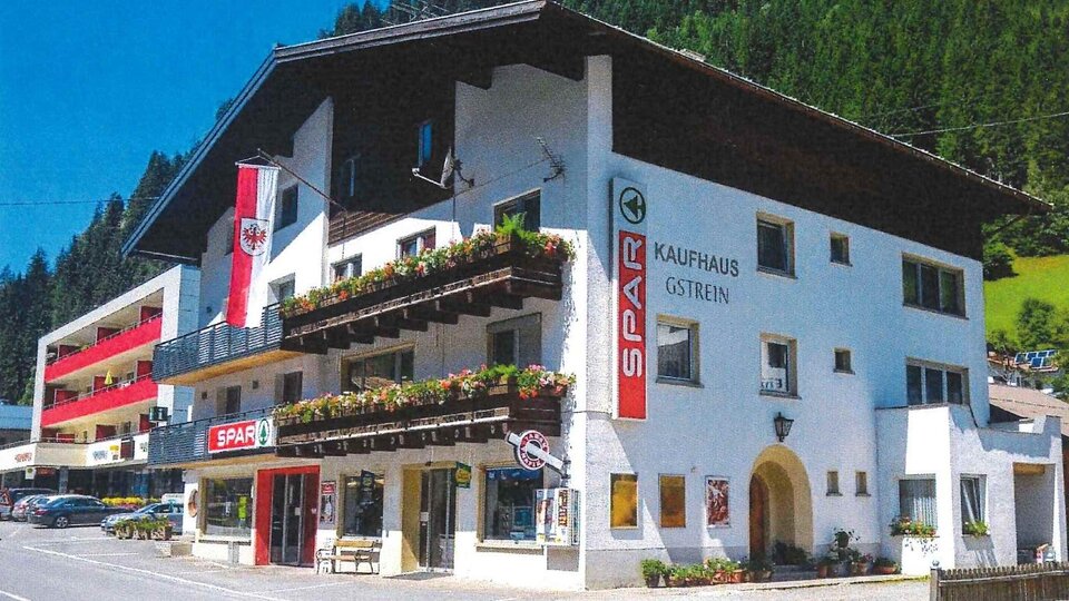 Ein kleines Geschäftshaus im alpenländischen Stil mit einem Spar-Supermarkt im Erdgeschoss. Die Fassade ist mit Blumenbalkonen und Fahnen dekoriert, umgeben von grünen Bergen.
