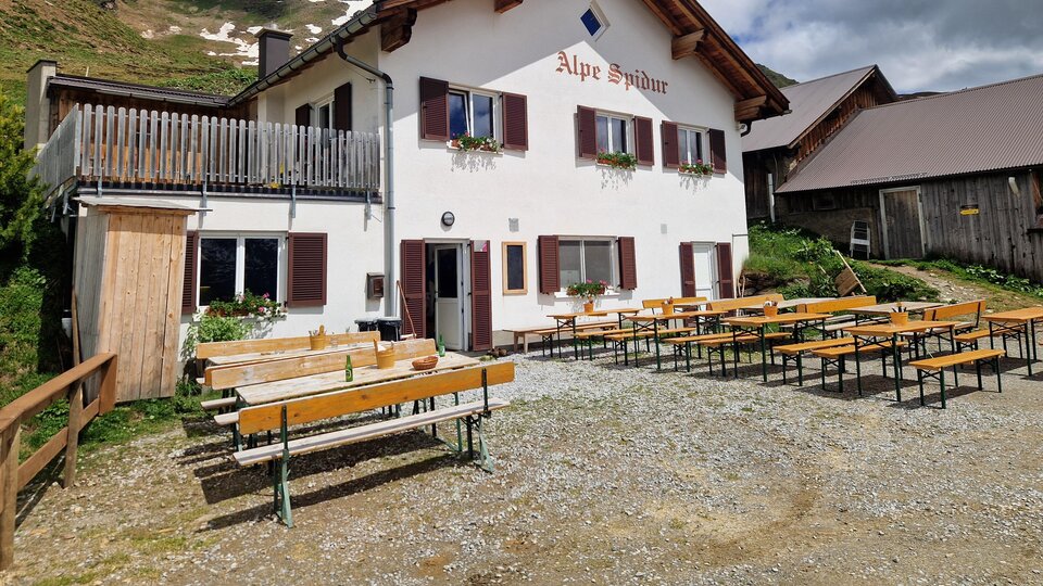 A cozy alpine house named "Alpe Spina". Outside there are many benches and tables in a picturesque mountain landscape.