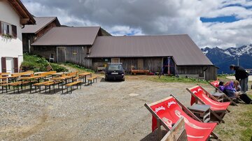 A rustic mountain inn with a large terrace. In the foreground, there are deck chairs and wooden tables, while in the background, the mountains and clouds are visible.
