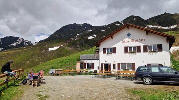 A cozy alpine house surrounded by tall mountains. There are some seating areas and cars visible on the premises.