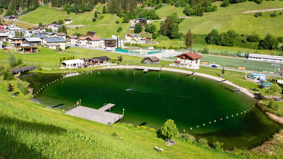 A picturesque pond surrounded by green meadows and mountains. In the background, several houses and facilities are visible.