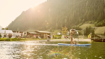 A person is standing on a stand-up paddleboard on a calm lake. In the background, there are mountains and a cozy building, while the sun is shining.