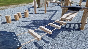 A playground bridge made of wood with ropes, leading over a gravel area. In the background, additional play equipment and a green area are visible.