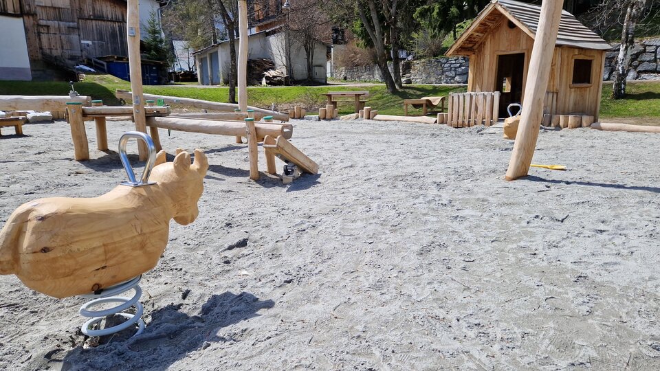A playground with a wooden rocking horse and a playhouse. The ground is made of sand and there are other play equipment nearby.