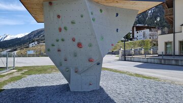 A modern outdoor climbing wall with colorful grips. It stands on a gravel area in front of a scenic backdrop.