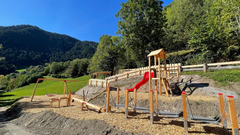 A playground in nature with wooden elements and a slide. In the background, green hills and trees can be seen.