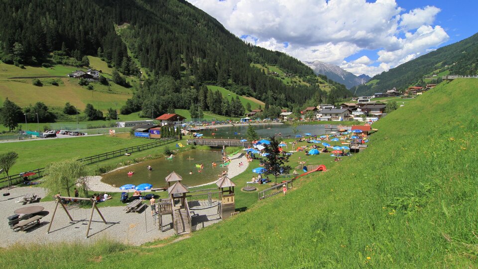 A picturesque park with a swimming lake and green meadows. In the background, mountains and houses can be seen, while people relax and enjoy the sun.